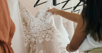 A person holding up a white lace wedding dress on a hanger, examining its floral details in a bridal shop.