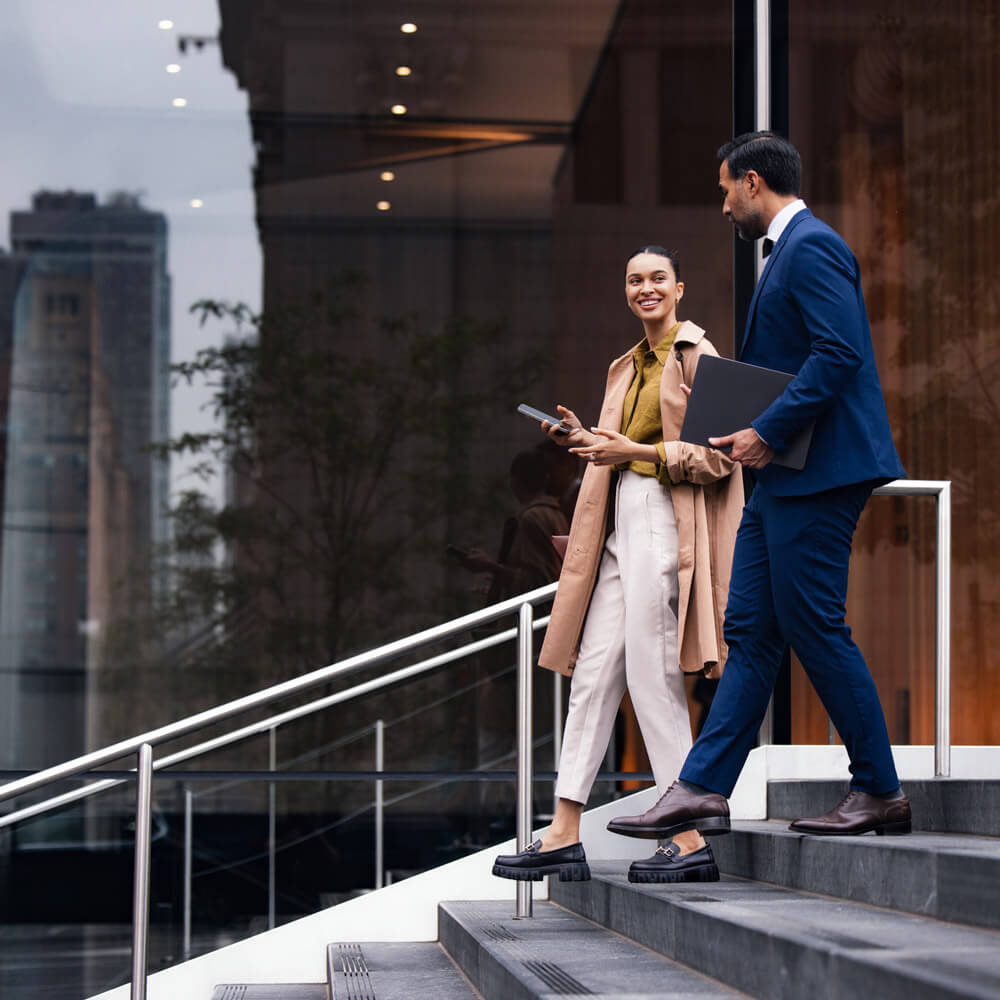 Two business professionals walk down the steps outside a modern office building while talking, one holding a laptop and the other a phone.