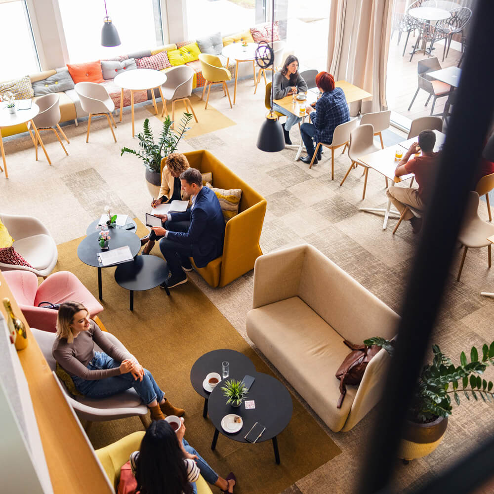 People sit and work or chat in a bright modern café-style coworking space with sofas, small tables, and large windows.