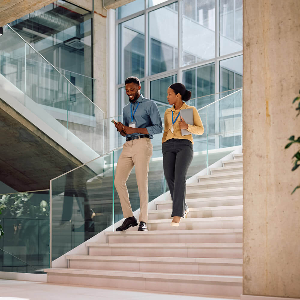 Two professionals wearing ID badges walk down a modern office staircase while talking, one holding a phone and the other carrying a tablet.